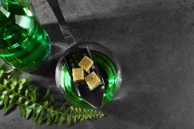 Photo of Absinthe in glass, spoon, brown sugar cubes, and fern leaf on gray textured table, above view with space for text. Alcoholic drink