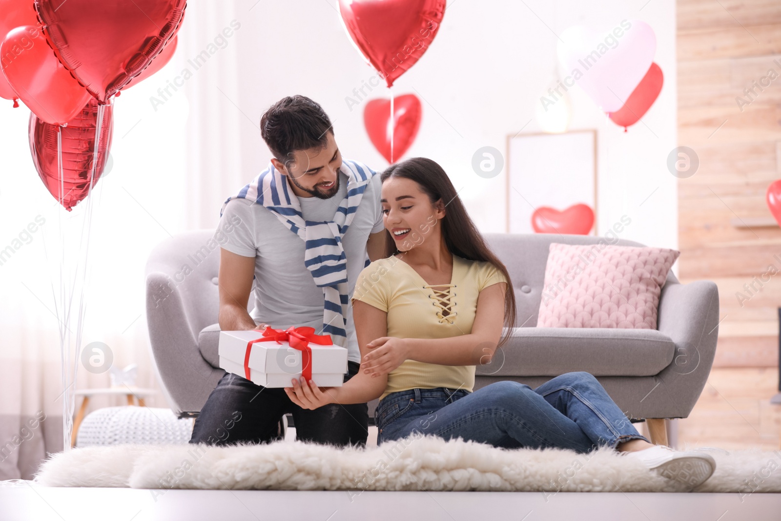 Photo of Young man presenting gift to his girlfriend in living room decorated with heart shaped balloons. Valentine's day celebration