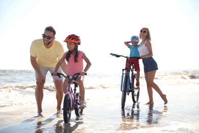 Photo of Happy parents teaching children to ride bicycles on sandy beach near sea