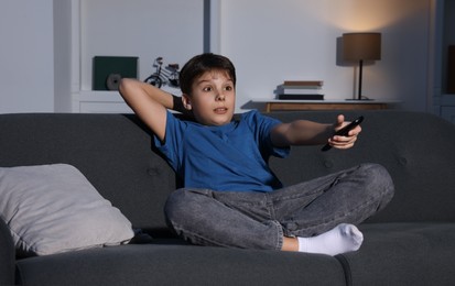 Emotional boy changing TV channels with remote control on sofa at home