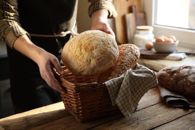 Photo of Man holding wicker basket with different types of bread at wooden table indoors, closeup