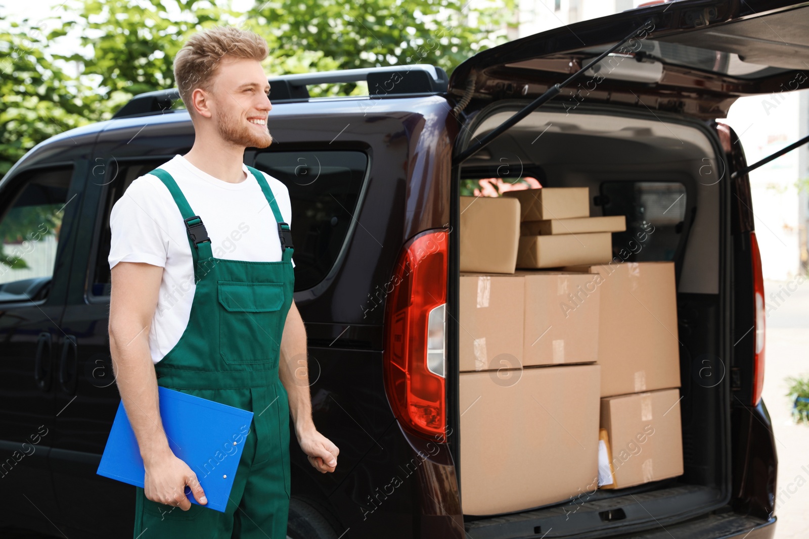 Photo of Young courier holding clipboard near delivery car with parcels outdoors