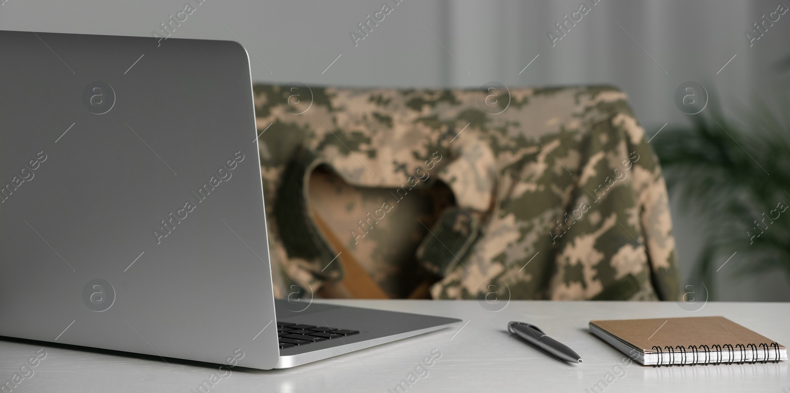 Image of Military education. Notebook, laptop and pen on white table. Chair with soldier's jacket indoors