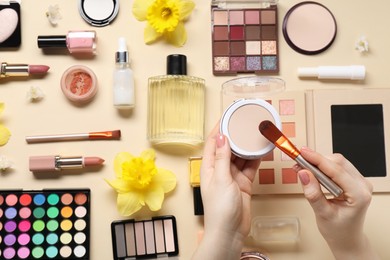 Photo of Woman applying face powder on brush, closeup. Different makeup products and beautiful spring flowers on beige background, top view