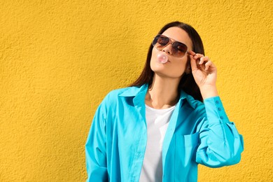Beautiful woman with sunglasses and blowing gum against orange wall, space for text