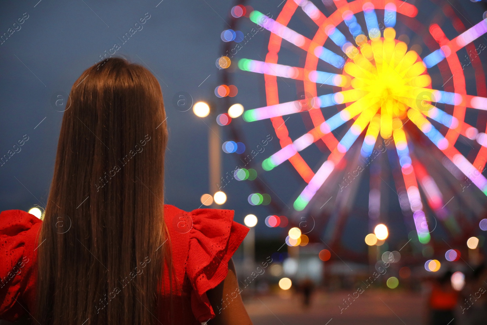 Photo of Beautiful young woman against glowing Ferris wheel in amusement park, back view. Space for text