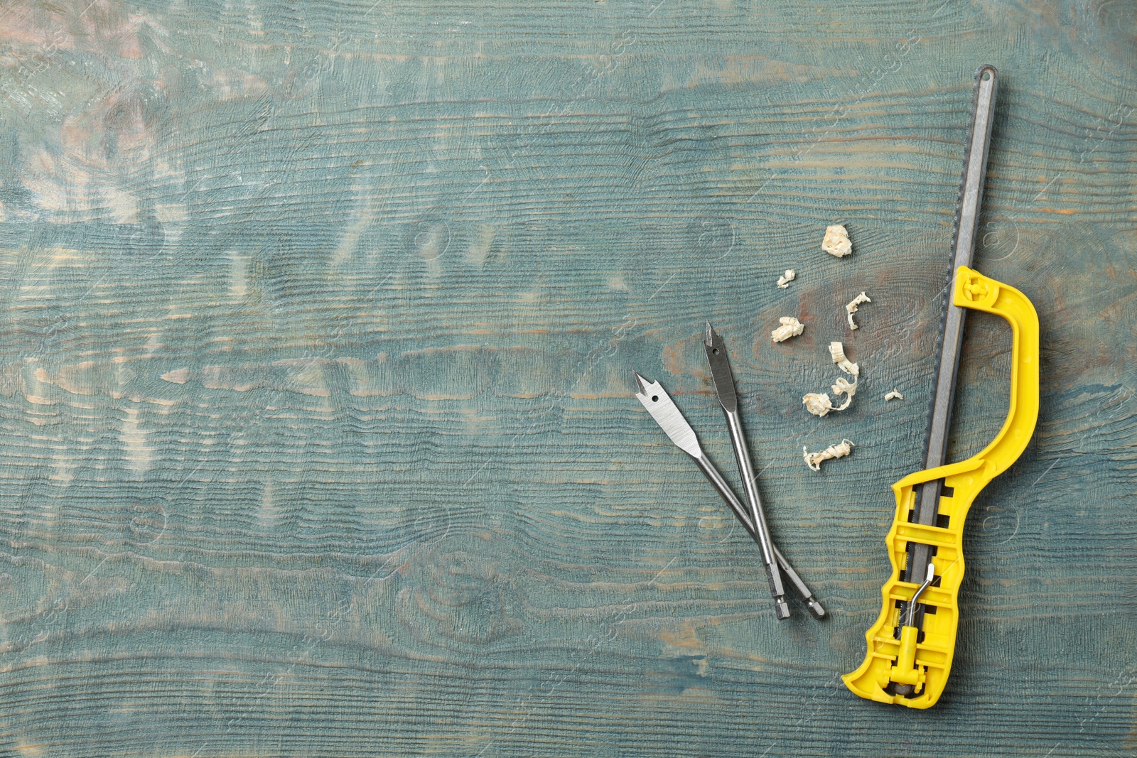 Photo of Small hand saw, spade drill bits and shavings on blue wooden background, flat lay with space for text. Carpenter's tools