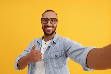 Smiling young man taking selfie and showing thumbs up on yellow background