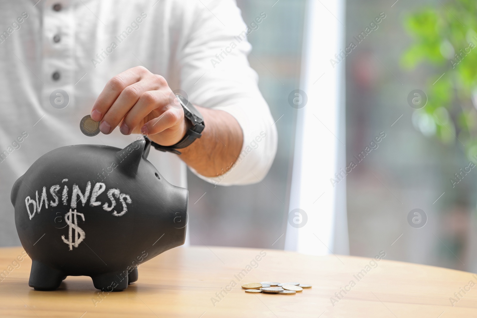Photo of Man putting money into piggy bank with word BUSINESS at table, closeup. Space for text
