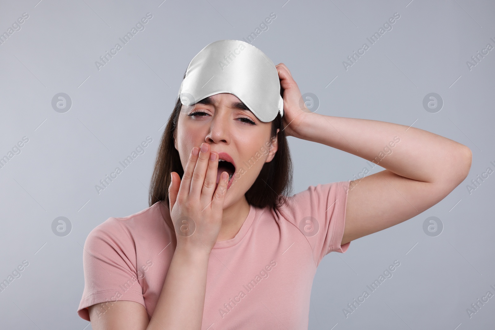 Photo of Tired young woman with sleep mask yawning on light grey background. Insomnia problem