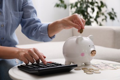 Woman with calculator putting coin into piggy bank at table indoors, closeup