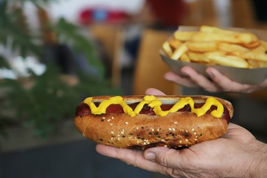 Woman holding fresh tasty hot dog with mustard and french fries in cafe, closeup. Space for text