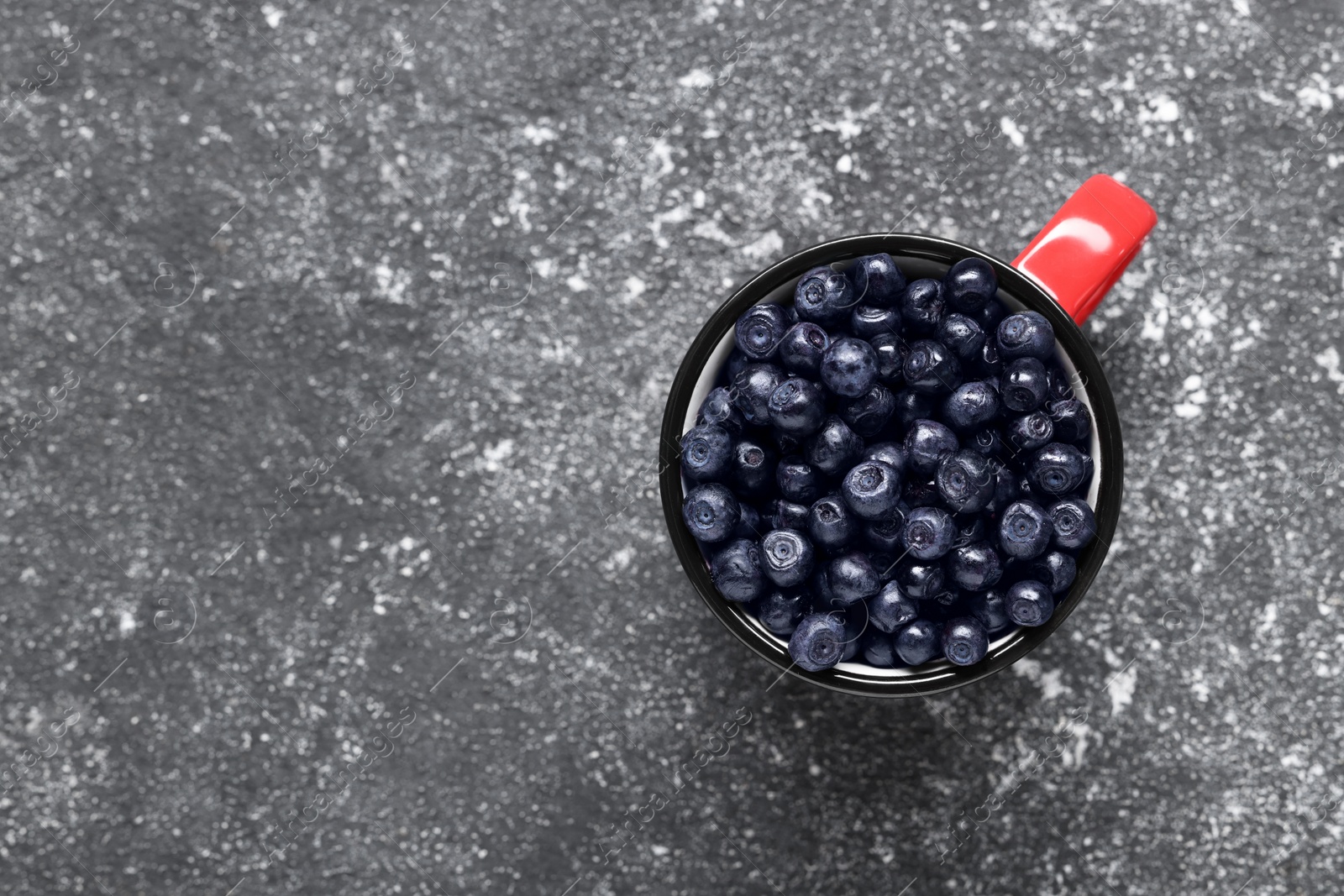 Photo of Ripe bilberries in mug on grey textured table, top view. Space for text