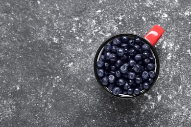 Ripe bilberries in mug on grey textured table, top view. Space for text