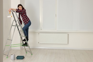 Photo of Young beautiful woman with roller on metal stepladder indoors. Room renovation