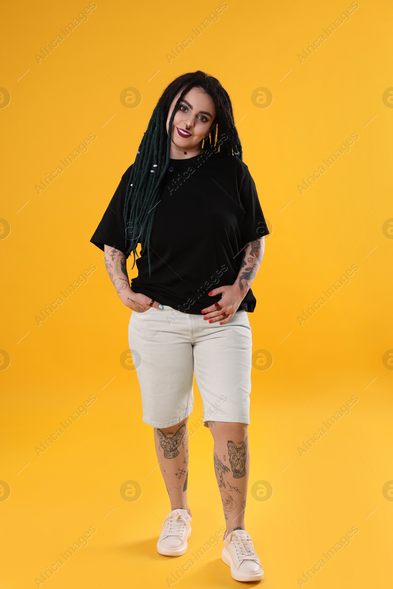 Photo of Beautiful young woman with tattoos on body, nose piercing and dreadlocks against yellow background