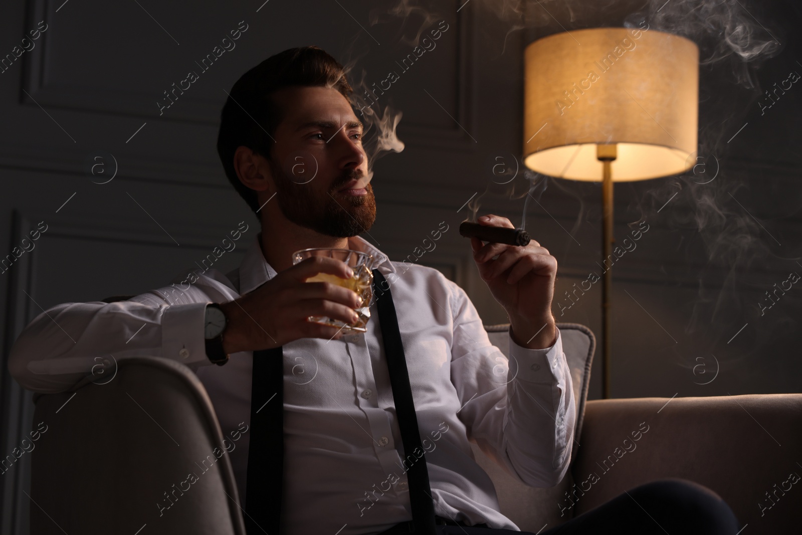 Photo of Handsome man with glass of whiskey smoking cigar at home