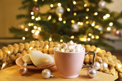 Photo of Cup of delicious cocoa, cookies and decor on wooden table against blurred Christmas tree with lights