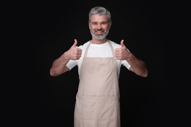 Photo of Happy man in kitchen apron showing thumbs up on black background. Mockup for design