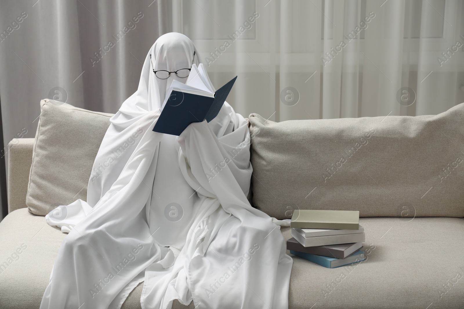 Photo of Creepy ghost. Person covered with white sheet reading book on sofa at home