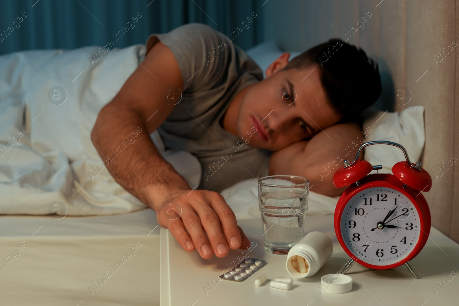 Photo of Man suffering from insomnia in bed at home, focus on pills and alarm clock