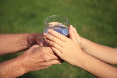 Photo of Child giving glass of water to elderly woman outdoors on sunny day, closeup