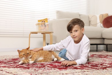 Photo of Little boy petting cute ginger cat on carpet at home