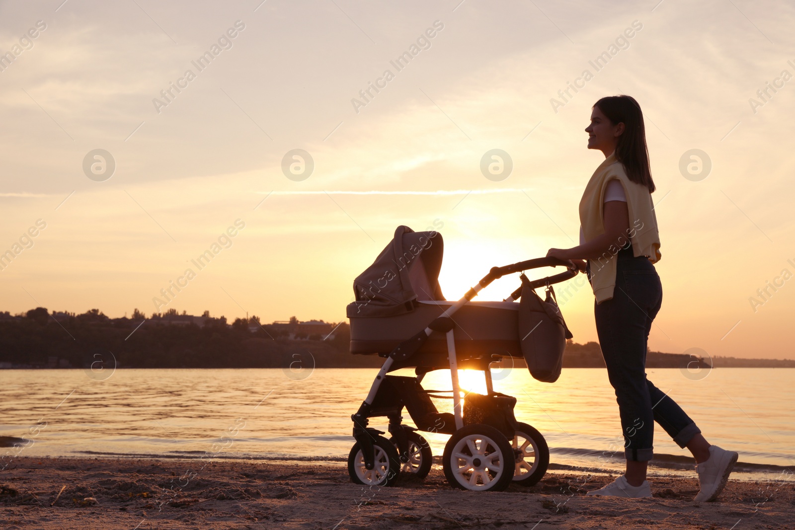 Photo of Happy mother with baby in stroller walking near river at sunset