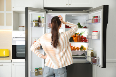 Photo of Young woman near open refrigerator in kitchen, back view