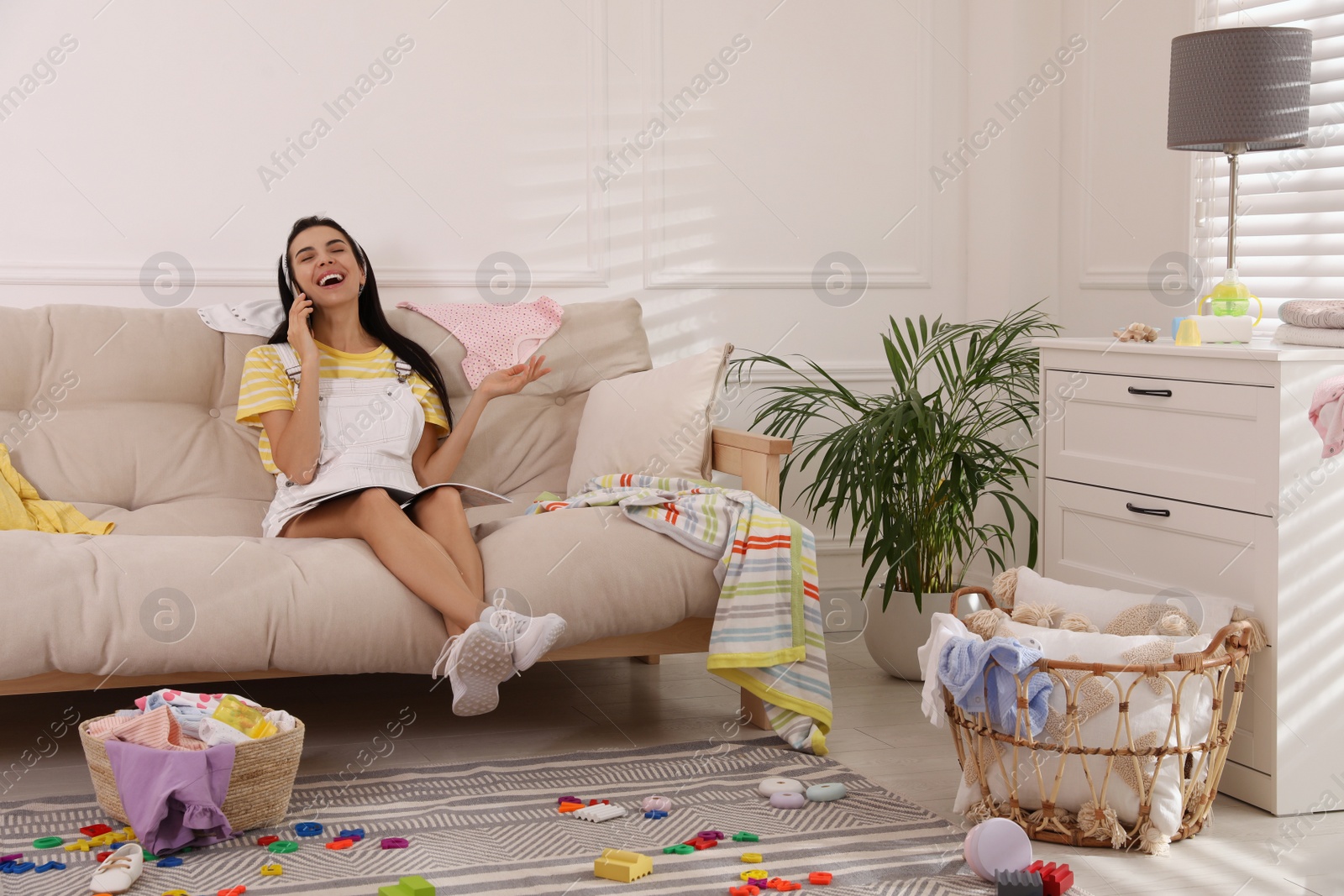 Photo of Young mother with magazine talking by mobile phone on sofa in messy living room