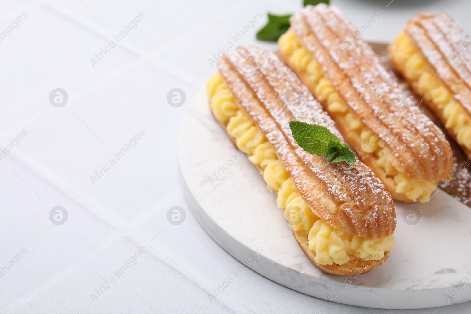 Photo of Delicious eclairs filled with cream and mint on white tiled table, space for text