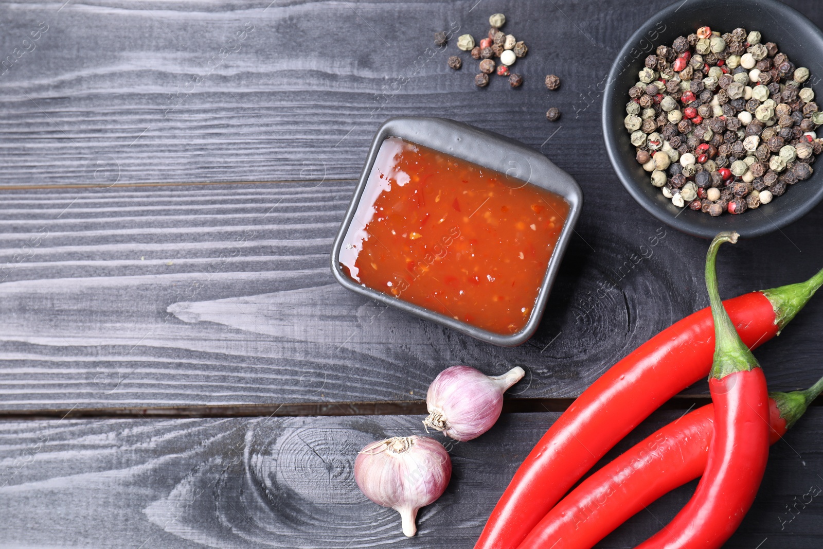 Photo of Spicy chili sauce, garlic, peppers and peppercorns on black wooden table, flat lay. Space for text