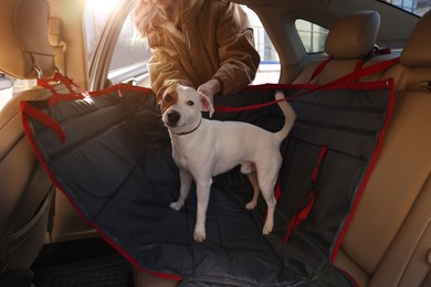 Photo of Woman fastening her cute Jack Russel Terrier dog with safety belt in bag carrier inside car. Pet accessory