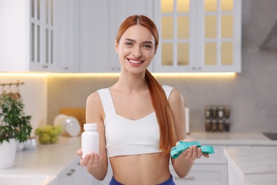 Photo of Happy young woman with bottle of pills and measuring tape in kitchen. Weight loss