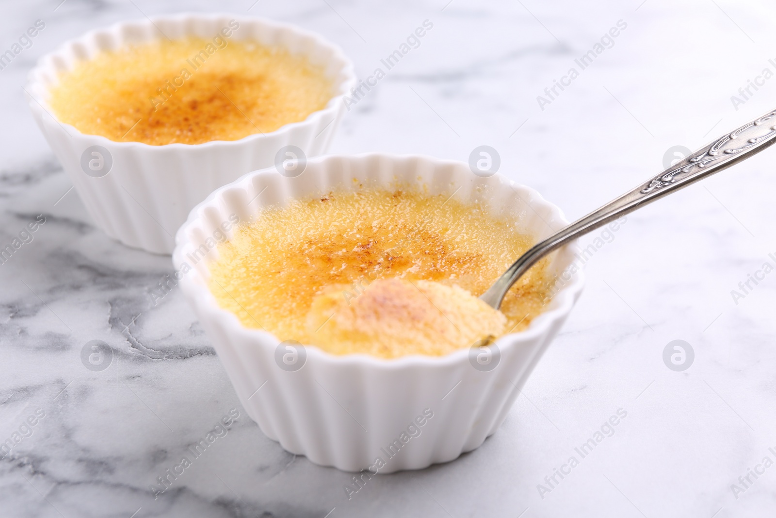 Photo of Delicious creme brulee in bowls and spoon on white marble table, closeup