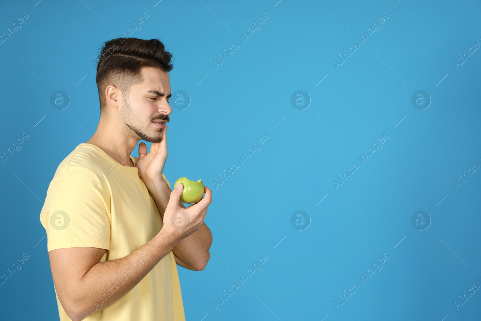 Photo of Emotional young man with sensitive teeth and apple on color background. Space for text