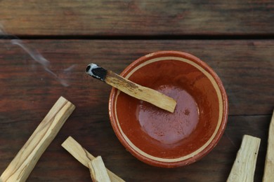 Photo of Palo santo stick smoldering in bowl on wooden table, flat lay