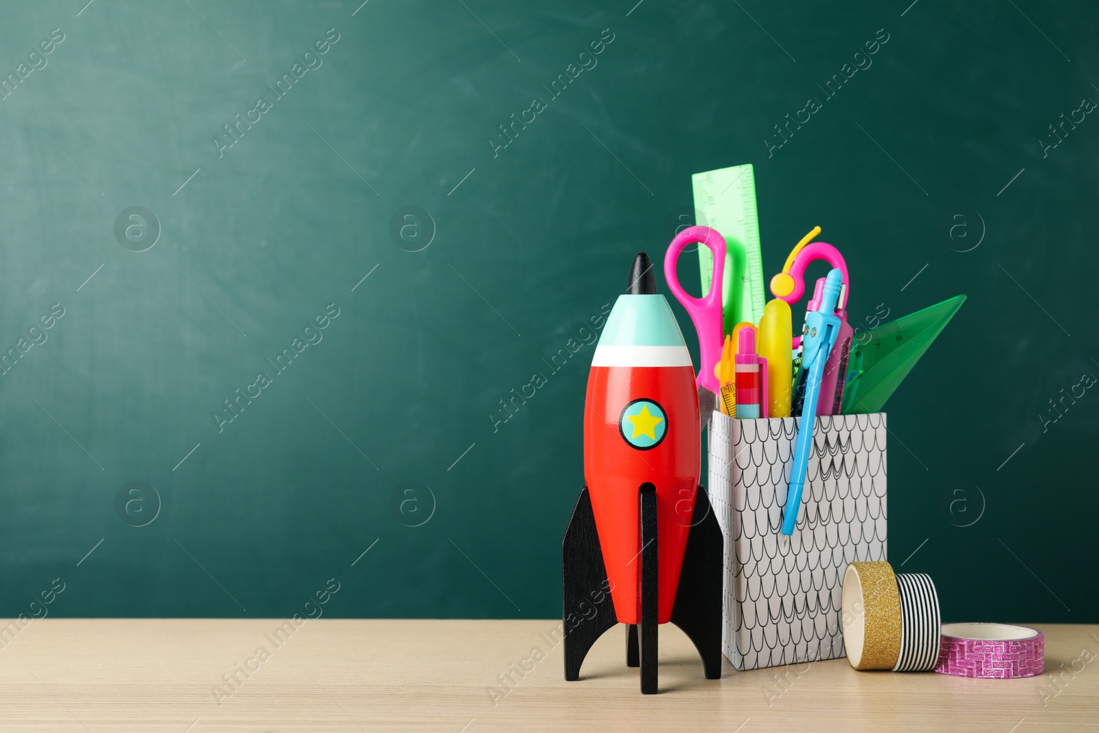 Photo of Bright toy rocket and school supplies on wooden desk. Space for text