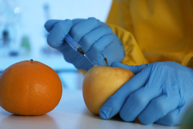 Photo of Scientist in chemical protective suit injecting apple at table, closeup