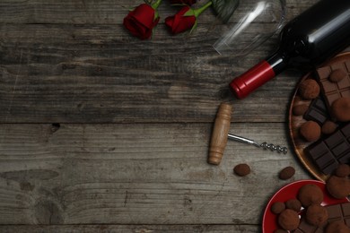 Photo of Chocolate sweets, bottle of red wine, glass, roses and corkscrew on wooden table, flat lay. Space for text