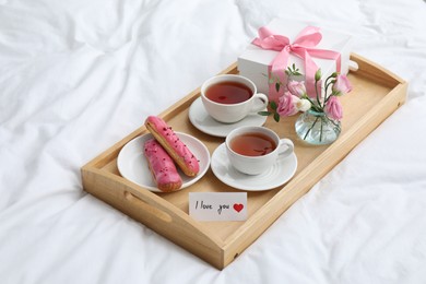 Photo of Tasty breakfast served in bed. Delicious eclairs, tea, gift box, flowers and card with phrase I Love You on tray