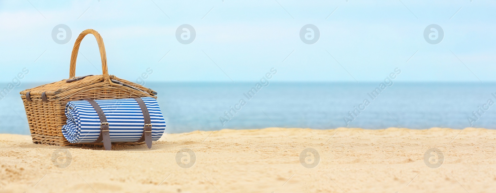 Image of Wicker picnic basket with blanket on sand near sea, space for text. Banner design