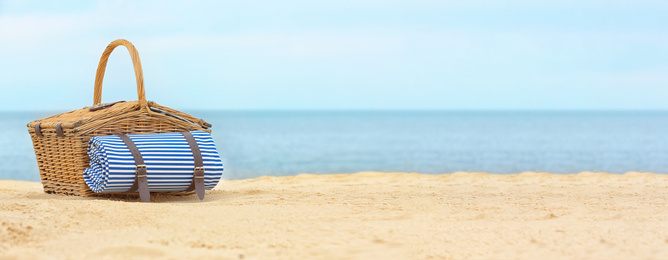 Image of Wicker picnic basket with blanket on sand near sea, space for text. Banner design