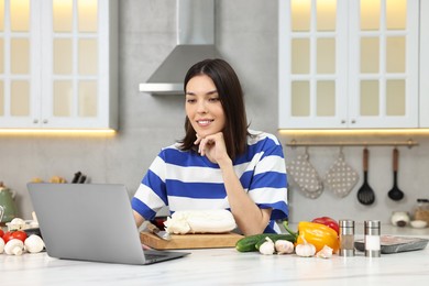 Photo of Young woman cooking while watching online course via laptop in kitchen