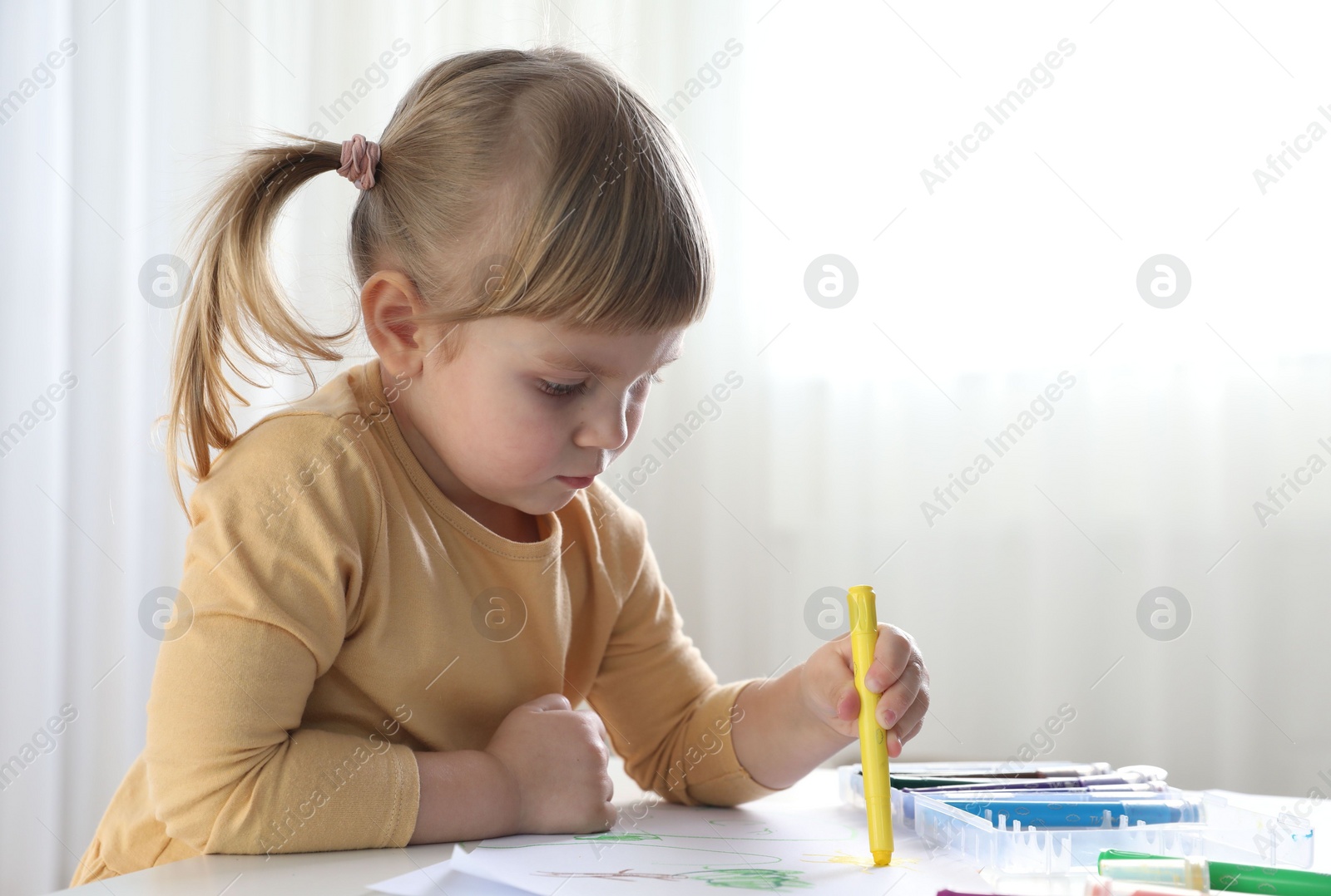 Photo of Cute little girl drawing with marker at white table indoors. Child`s art