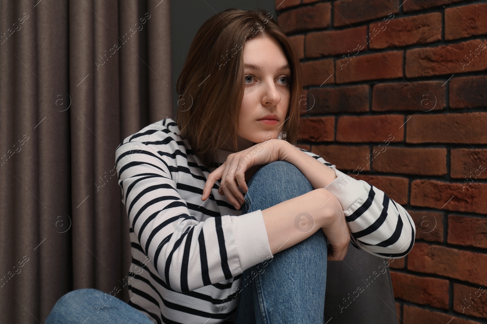 Photo of Sad young woman sitting on chair near brick wall indoors