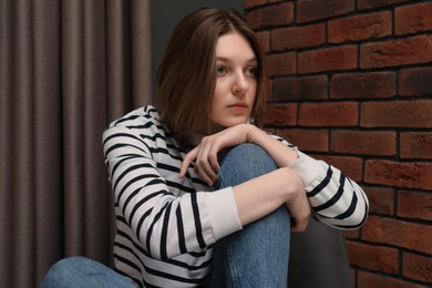 Photo of Sad young woman sitting on chair near brick wall indoors