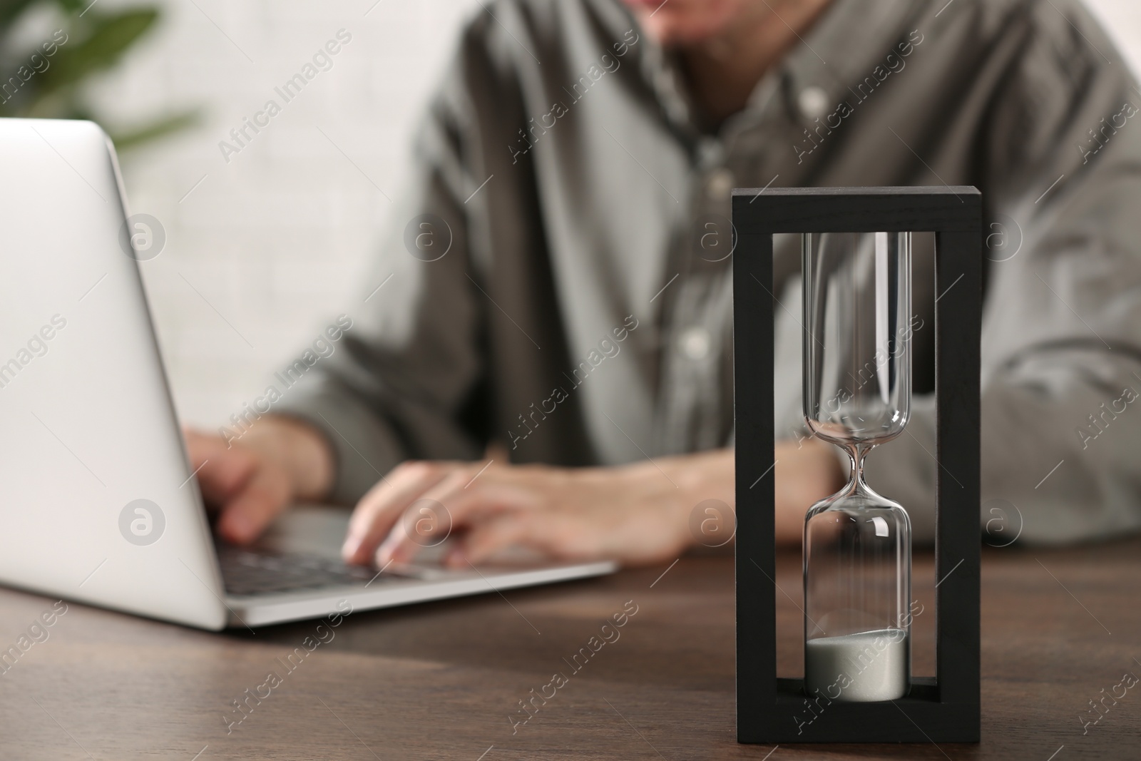 Photo of Hourglass with flowing sand on wooden table, selective focus. Man using laptop indoors