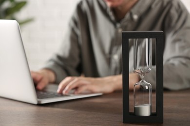 Photo of Hourglass with flowing sand on wooden table, selective focus. Man using laptop indoors