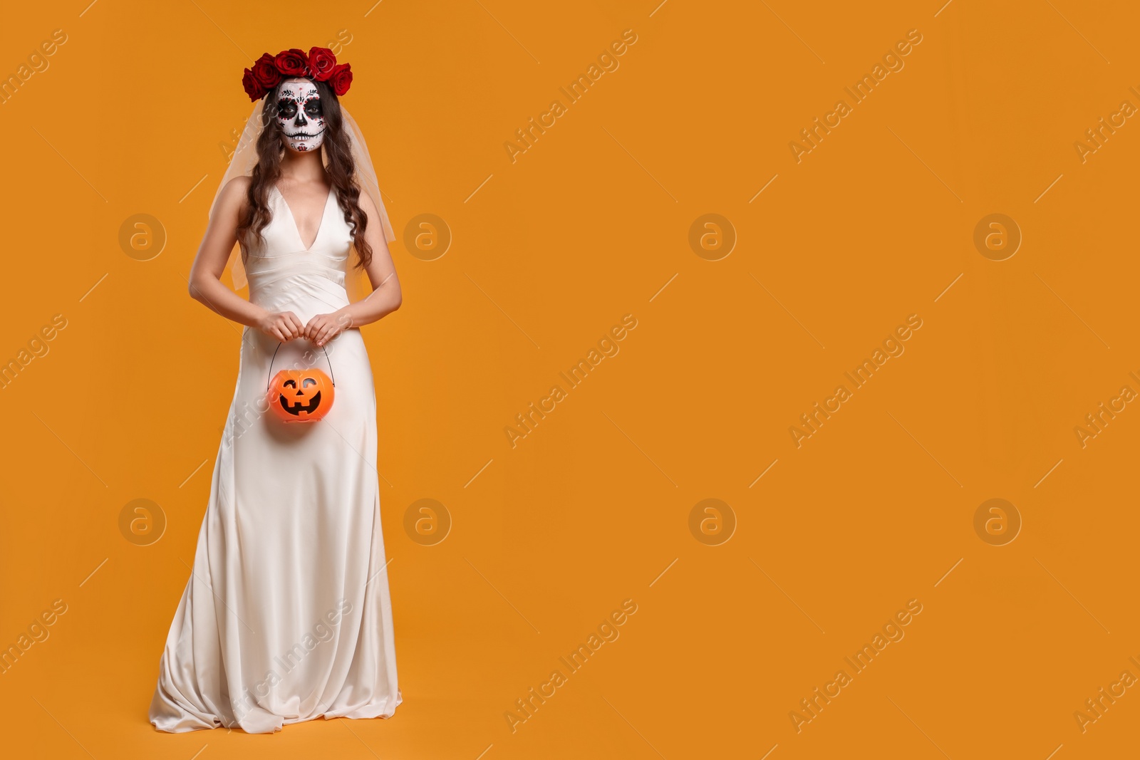 Photo of Young woman in scary bride costume with sugar skull makeup, flower crown and pumpkin bucket on orange background, space for text. Halloween celebration